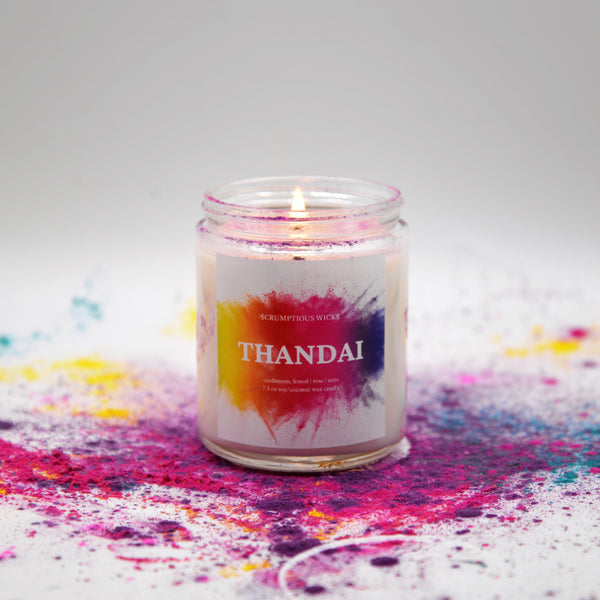 Thandai scented candle in glass jar with lit flame and vibrant, colorful label surrounded by colored powder for Holi, the festival of colors or festival of spring.